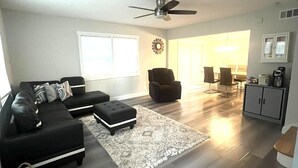 Una sala moderna con un sofá seccional negro, un pequeño otoman, un sillón oscuro y un comedor con mesa y sillas.