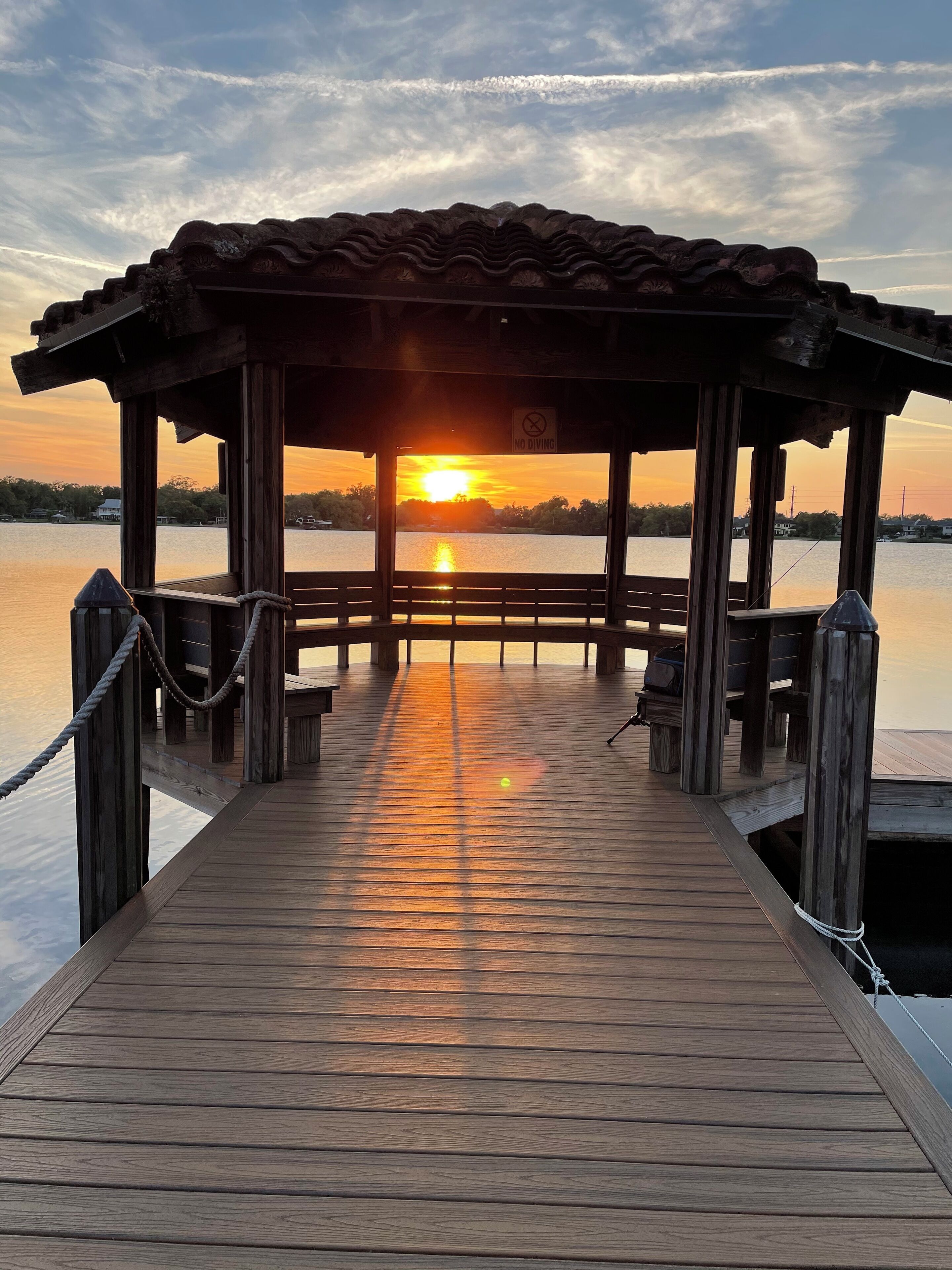A wooden pier leading to a covered area with a 'No Diving' sign, overlooking a calm body of water during sunset.