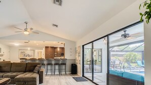 A spacious living room with a large sofa, a dining area with chairs, a kitchen in the background, and a sliding glass door leading to an outdoor patio. - Beautiful Cozy 3-bedroom House (Naples)