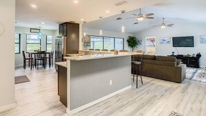 A modern kitchen with a central island, dining area with chairs, a living room with a sofa, and a TV. - Beautiful Cozy 3-bedroom House (Naples)