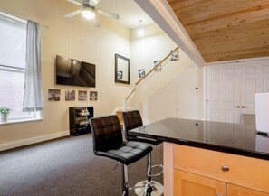 A room with a wooden ceiling, a ceiling fan, a TV mounted on the wall, a bar counter with two stools, and a staircase leading to another level.