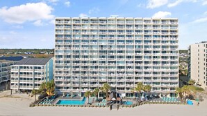 Ein mehrstöckiges Hotel mit zahlreichen Balkonen, einem Poolbereich inklusive Liegestühlen und Blick auf den Strand.