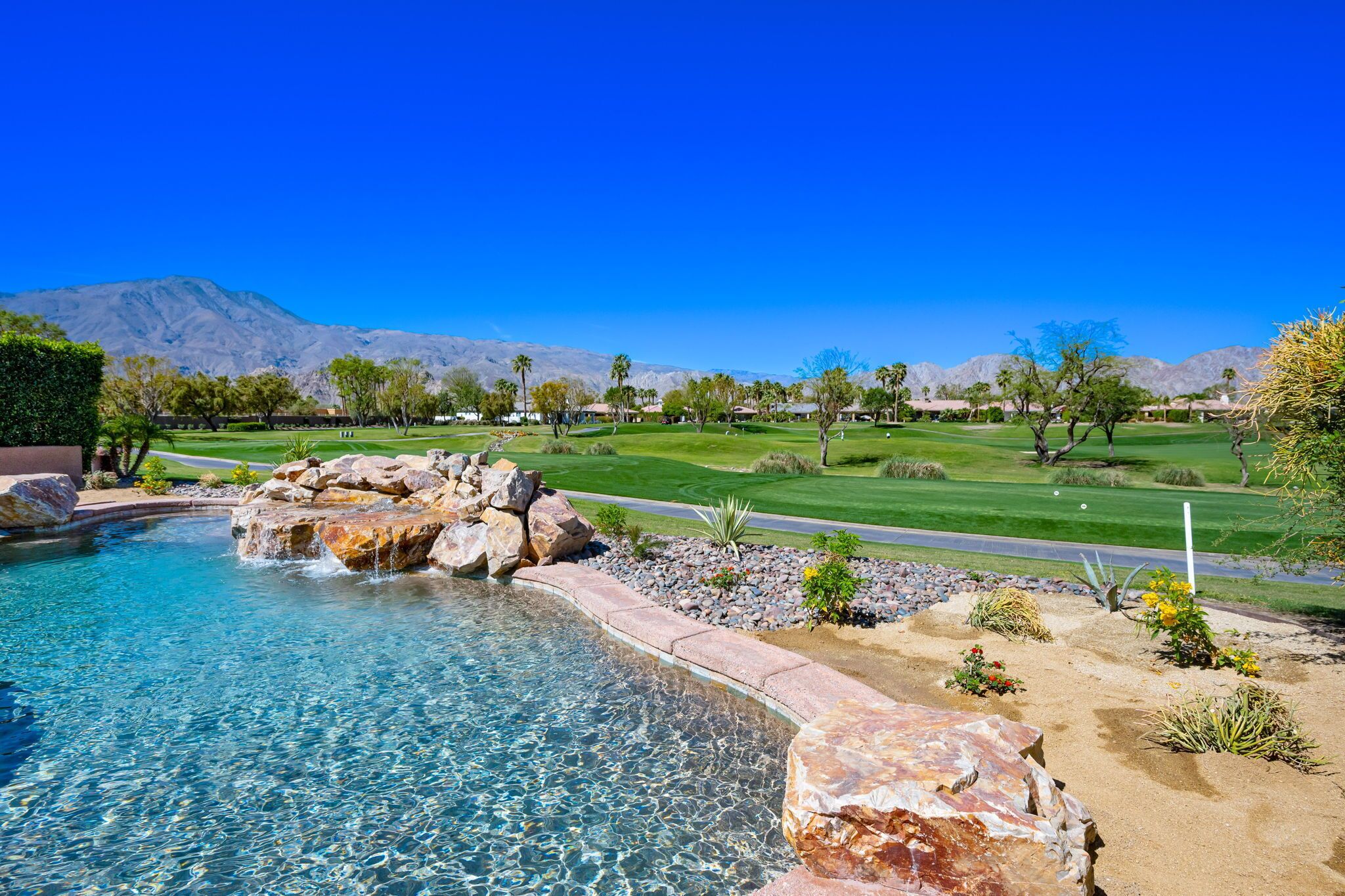 Una piscina cristalina con rocas, un campo de golf y montañas al fondo.