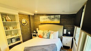 Een slaapkamer met een bed, nachtkastje, dressoir, bankje en raam met jaloezieën.
