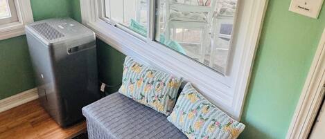 Ein Zimmer mit Fenster, eine Bank mit blumigen Kissen, ein Heizgerät und ein Lichtschalter mit der Aufschrift 'Porch light'.