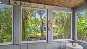 Een kamer met grote ramen die uitkijken op een tuin en een rivier, een houten bord boven de ramen, en een klein tafeltje met een mandje.