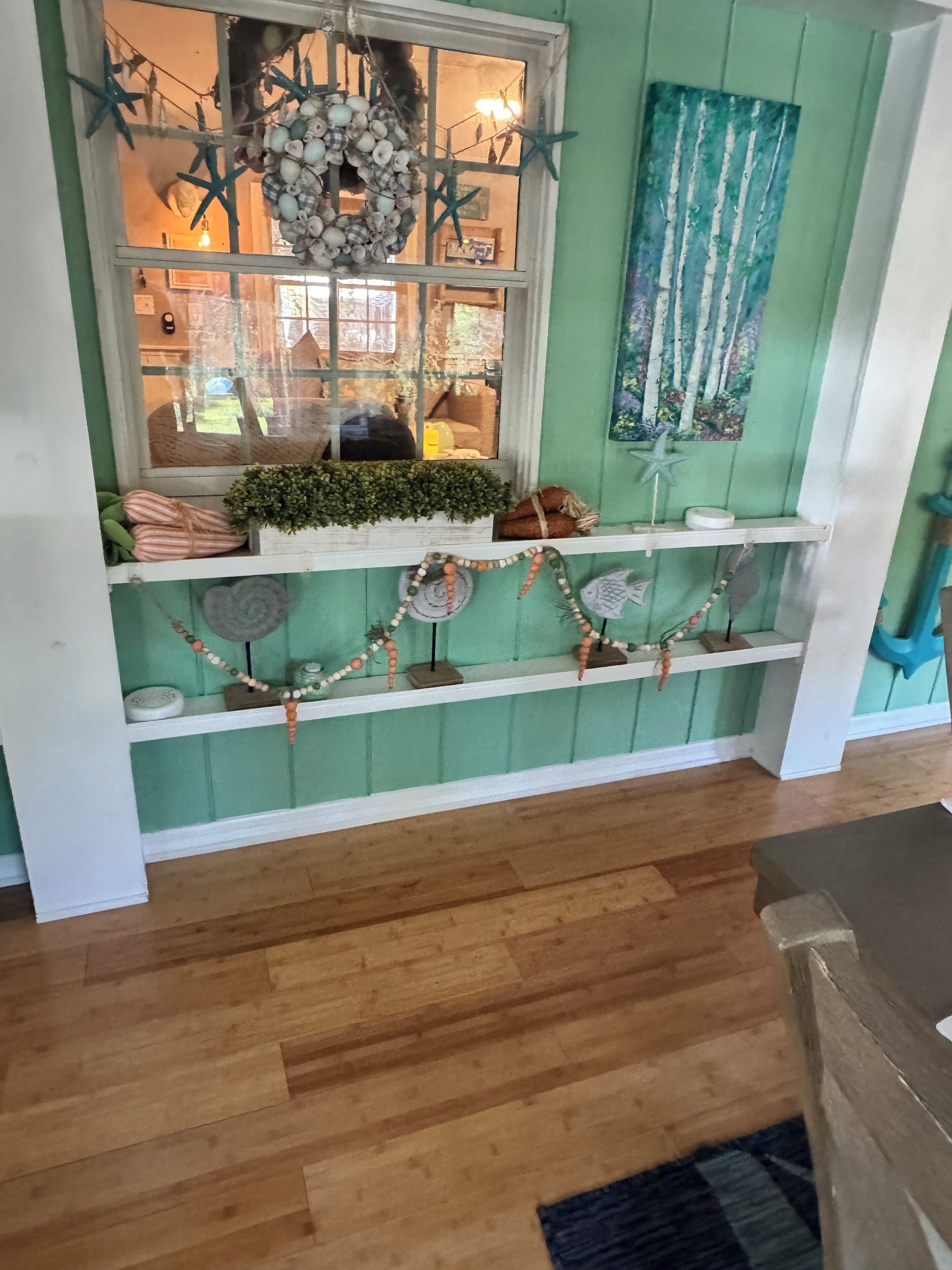 Ein Zimmer mit grüner Wand, einem Fenster mit Seestern-Dekorationen, einem Kranz und einem Baumgemälde.