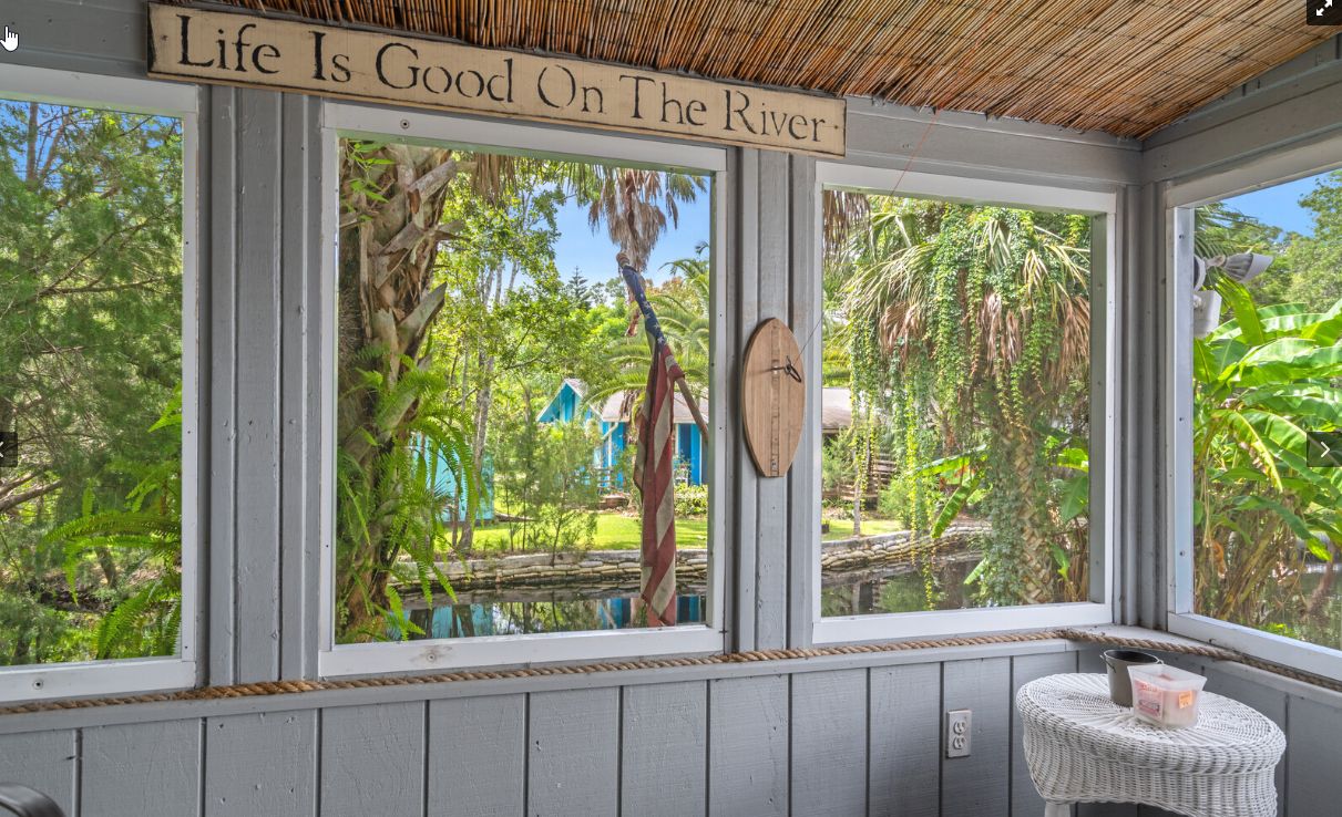 Ein Zimmer mit großen Fenstern, die auf einen Garten und einen Fluss blicken, ein Holzschild über den Fenstern und ein kleiner Tisch mit einem Korb.