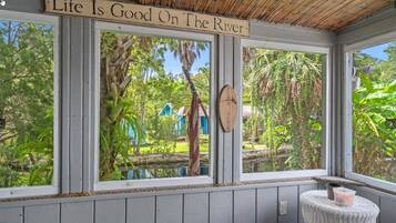 Ein Zimmer mit großen Fenstern, die auf einen Garten und einen Fluss blicken, ein Holzschild über den Fenstern und ein kleiner Tisch mit einem Korb.