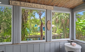 Ein Zimmer mit großen Fenstern, die auf einen Garten und einen Fluss blicken, ein Holzschild über den Fenstern und ein kleiner Tisch mit einem Korb.