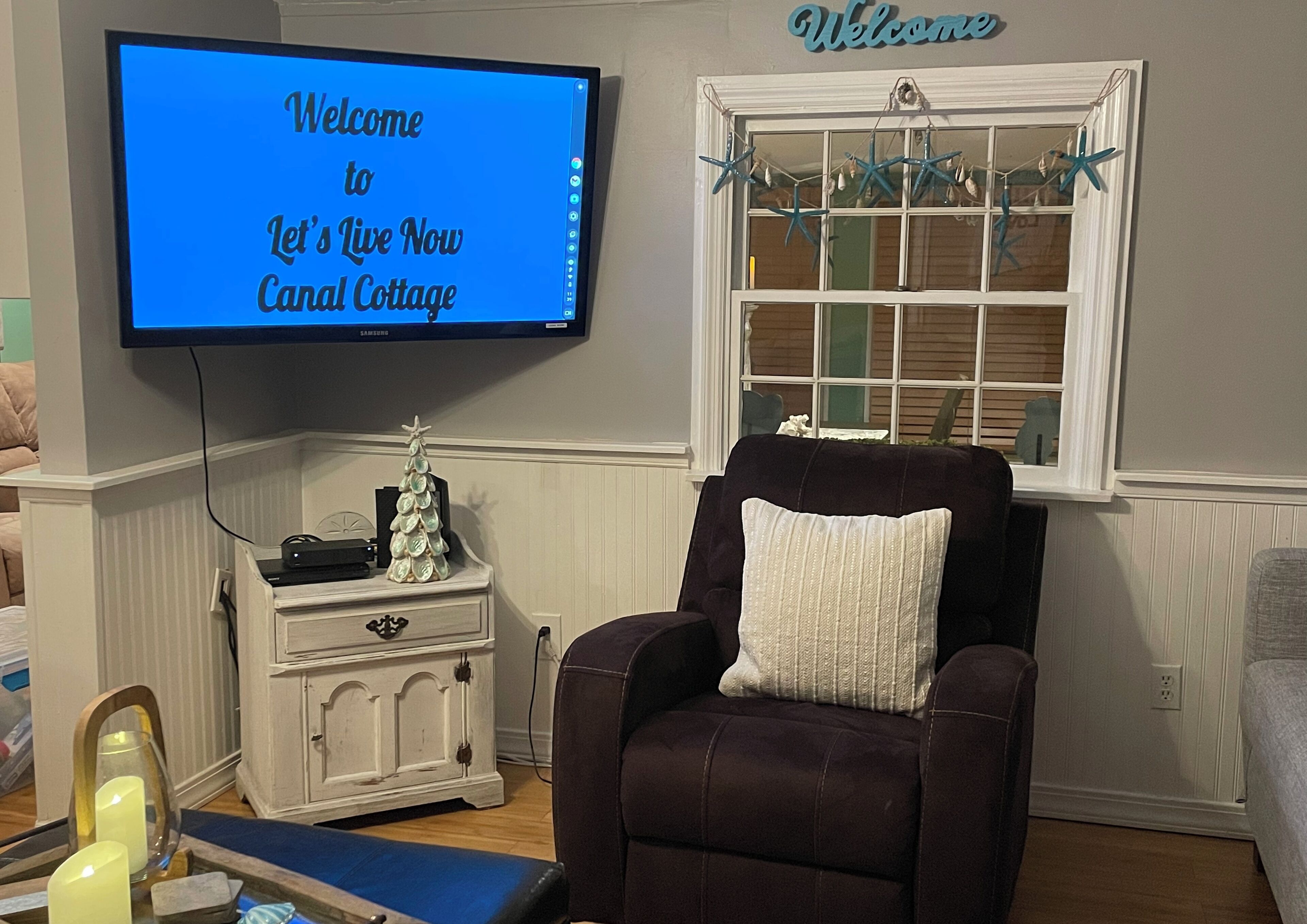A living room with a flat-screen TV displaying "Welcome to Let's Live Now Canal Cottage," a dark brown armchair with a white pillow, a small white cabinet with a decorative tree, and a window with starfish decorations.