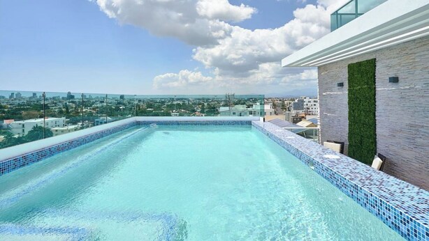 A rooftop infinity pool with a city view, surrounded by a modern building with a glass balcony and a vertical garden. - Modern & Cozy place w/pool & Parking in Downtown (Santo Domingo)