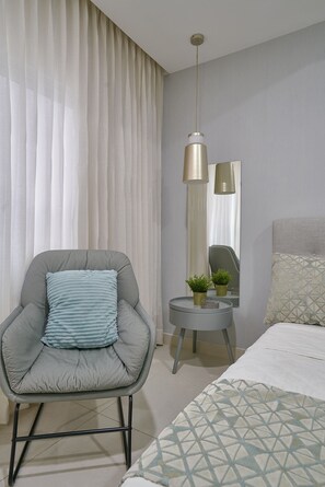 A bedroom with a grey armchair, a bedside table with a lamp, a bed with a patterned bedspread, and a large window with sheer curtains.
