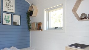 Une chambre à coucher comprenant un lit, une fenêtre, un bureau avec un ordinateur portable et des éléments de décoration murale, notamment des tableaux encadrés et une citation.