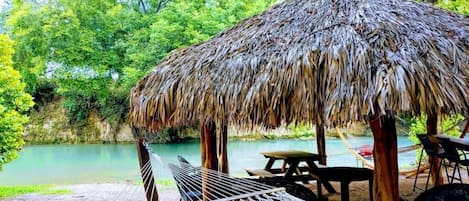 Un hamac, une structure à toit de chaume et une table de pique-nique près d’une étendue d’eau calme, avec une végétation luxuriante en arrière-plan.