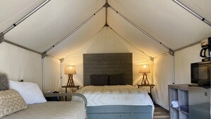 A tented accommodation with a bed, bedside lamps, a small kitchenette, and a television.