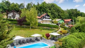A pool area with lounge chairs and umbrellas, surrounded by lush greenery and houses.