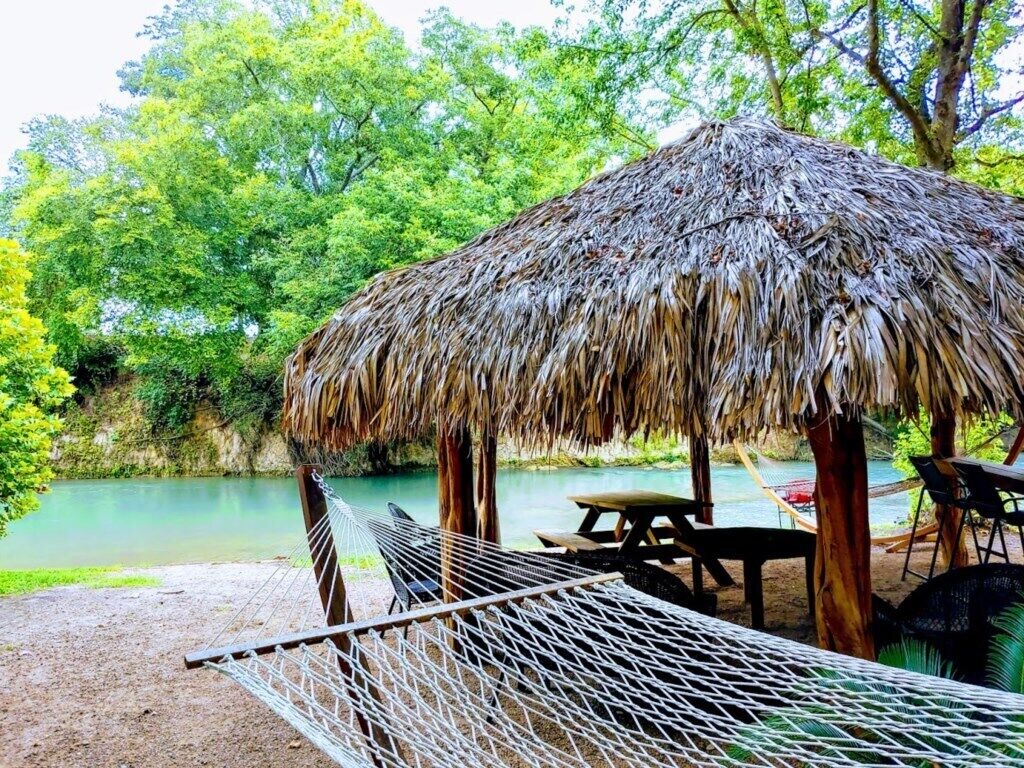Un hamac, une structure à toit de chaume et une table de pique-nique près d’une étendue d’eau calme, avec une végétation luxuriante en arrière-plan.