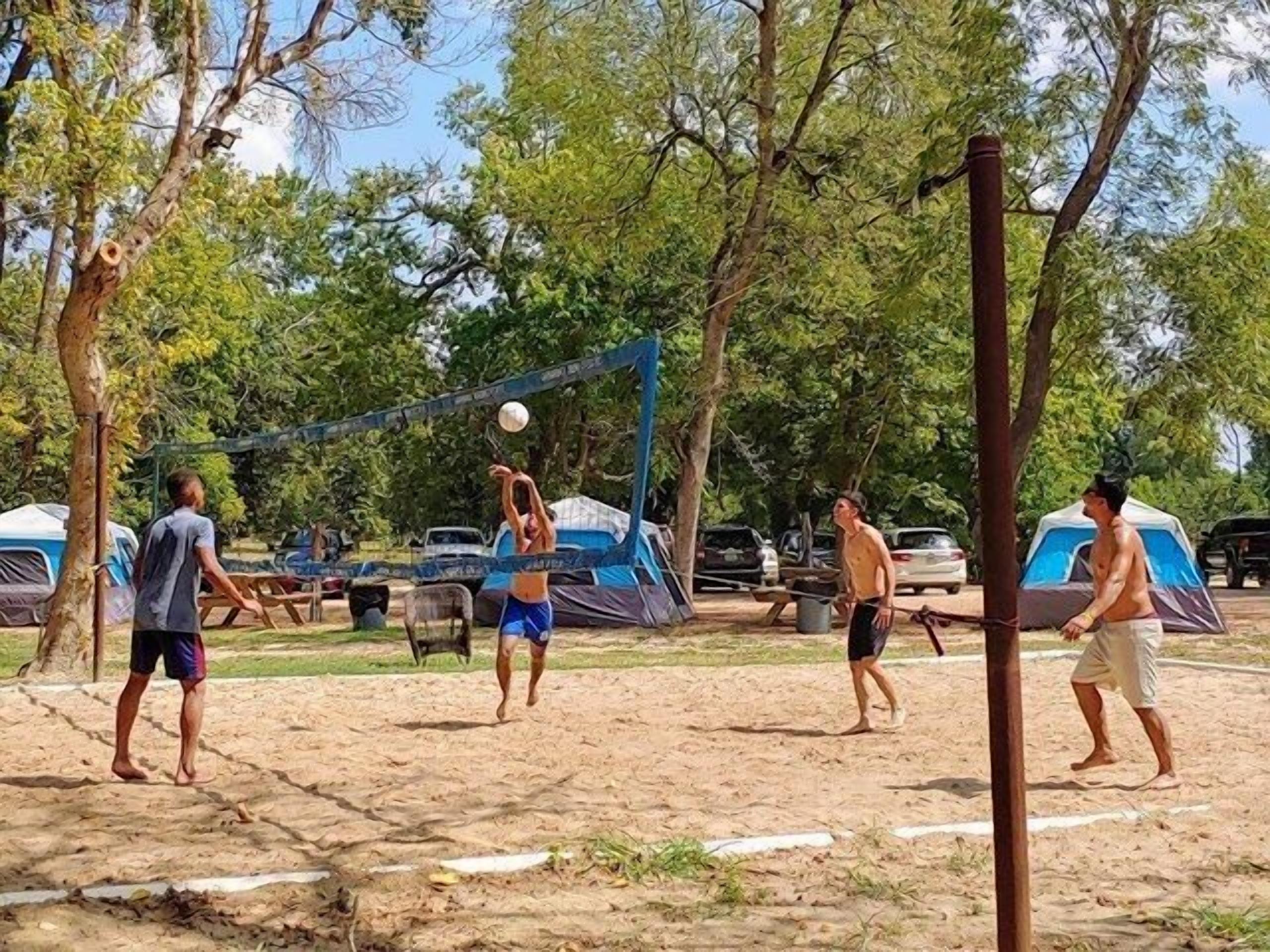 Una cancha de voleibol de arena, con tiendas de campaña y árboles al fondo.