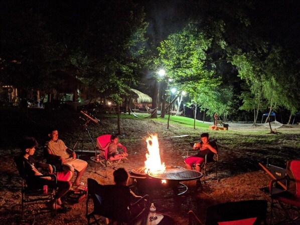 A group of people seated around a campfire in an outdoor setting with trees and a grassy area in the background.