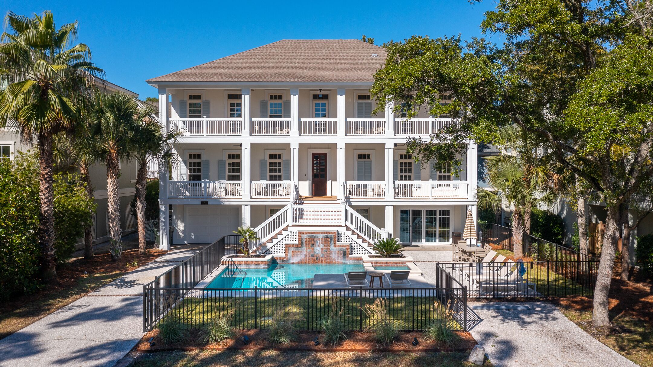 A two-story building with a swimming pool, surrounded by palm trees and a fenced patio area.