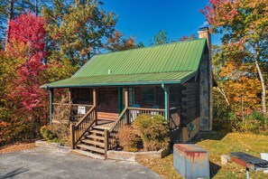 A rustic cabin with a green metal roof, wooden porch, and stone chimney.