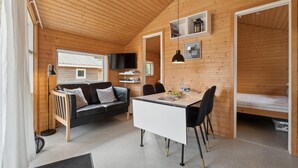 A cozy cabin interior with a wooden ceiling, a sofa, a dining table with chairs, a TV mounted on the wall, and a window providing natural light. - Beautiful home in Nexø with kitchen (Nexø)