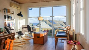 A spacious living area with a large window offering a view of the sea and buildings. There is a wooden table, a bookshelf, and a rocking chair.