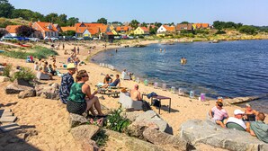 A beach with people relaxing on chairs and sunbathing, a clear blue sky, and houses in the background.