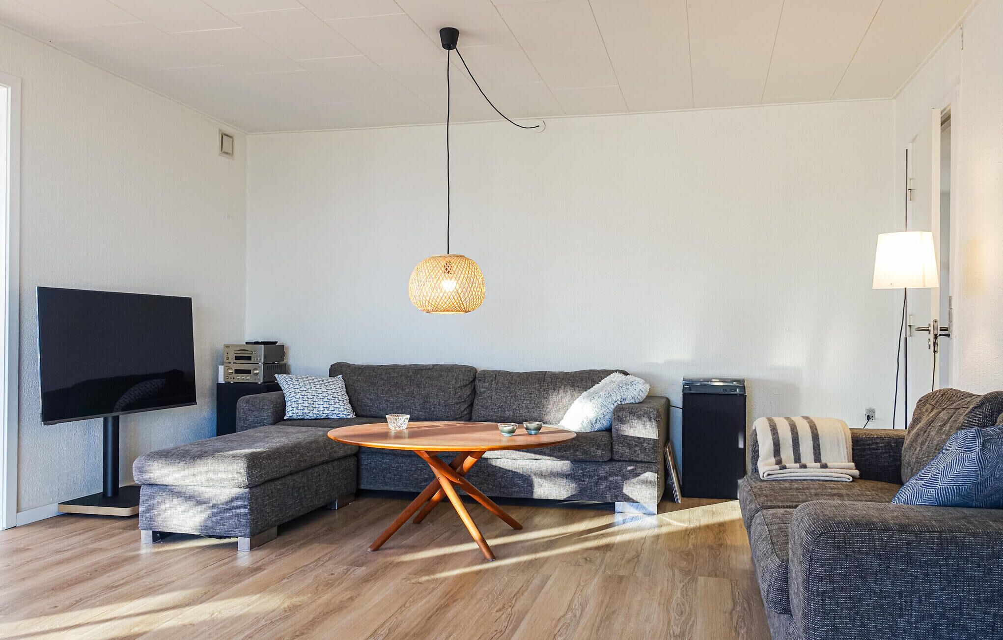 Un salon moderne avec un canapé d’angle, une table basse ronde en bois, un téléviseur à écran plat et une suspension décorative en fibres tressées.