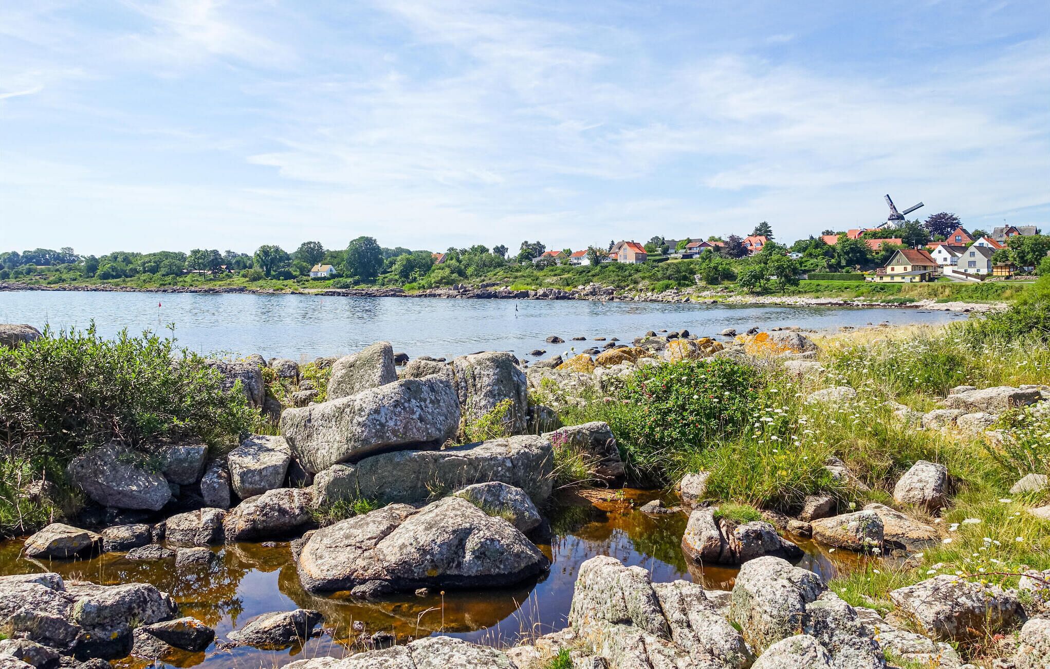 Paesaggio costiero con un litorale roccioso, un piccolo corso d'acqua e case sullo sfondo.