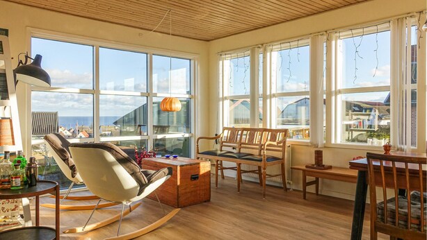 A spacious living room with large windows, a rocking chair, a wooden coffee table, and a desk with chairs.