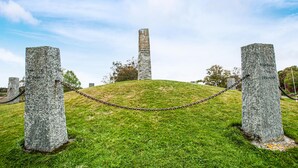 Monumento de piedra con inscripciones, rodeado de césped y árboles, con una barrera de cadena en primer plano.