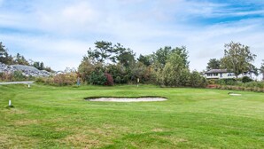 Un campo de golf con un búnker de arena, árboles y una casa al fondo.