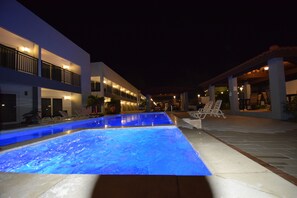 En swimmingpool med blå belysning, liggestole og en bygning med balkoner og lys.