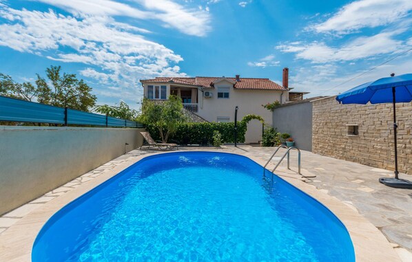 A swimming pool with a clear blue water, surrounded by a stone patio, a house with a tiled roof, and a blue umbrella.