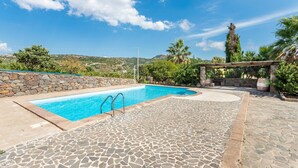 A rectangular swimming pool with a stone-tiled deck, surrounded by a stone wall and lush greenery, with a clear blue sky above. - Stunning home in Bosa (Bosa)