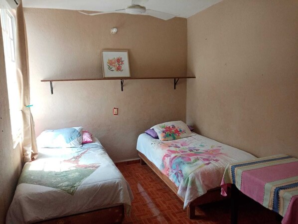 A room with two single beds, a small shelf with a floral picture, and a window with curtains.