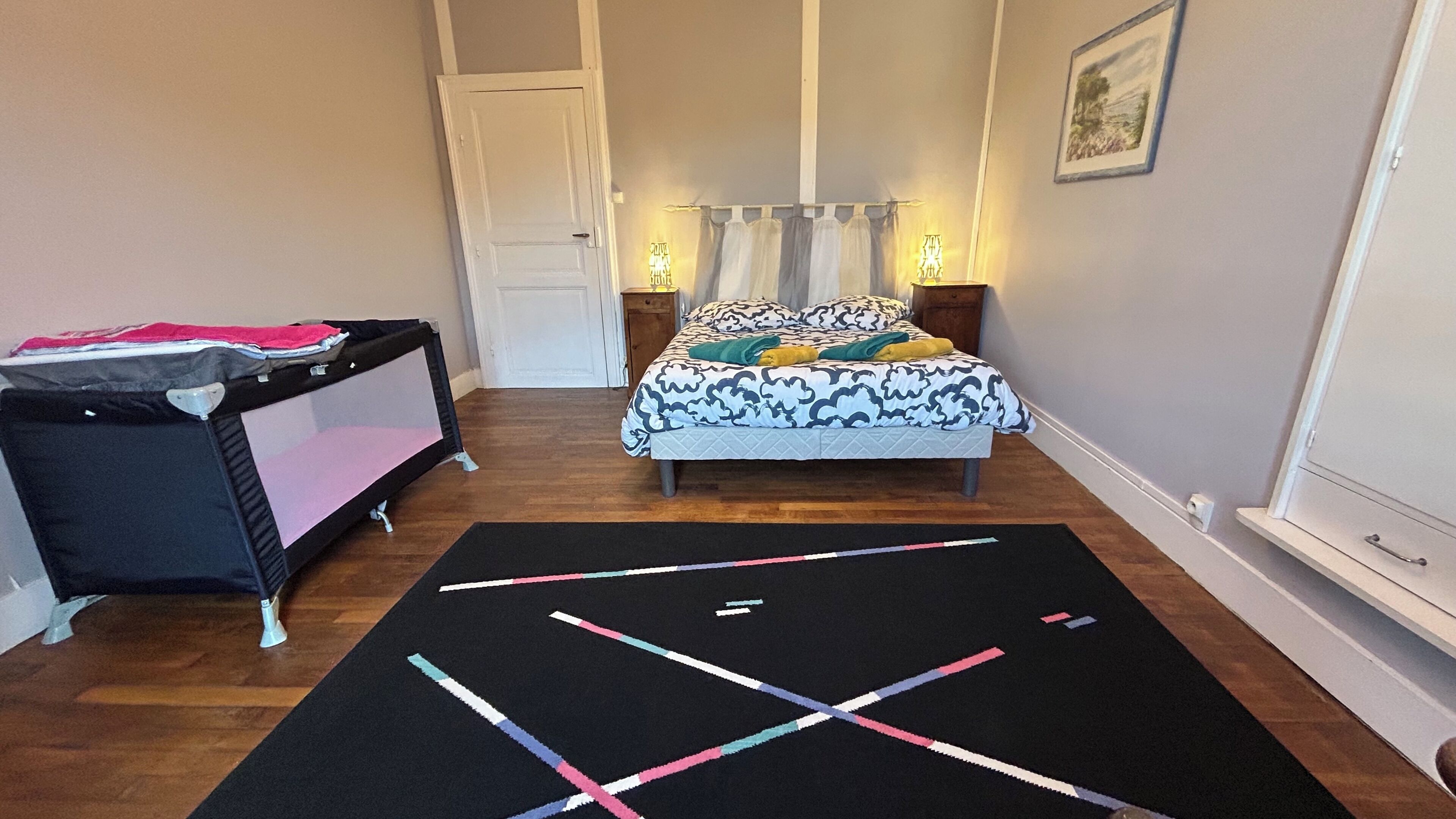 Ein Schlafzimmer mit einem Bett, einem Kinderbett, einem schwarzen Teppich mit geometrischen Mustern und einer Tür.
