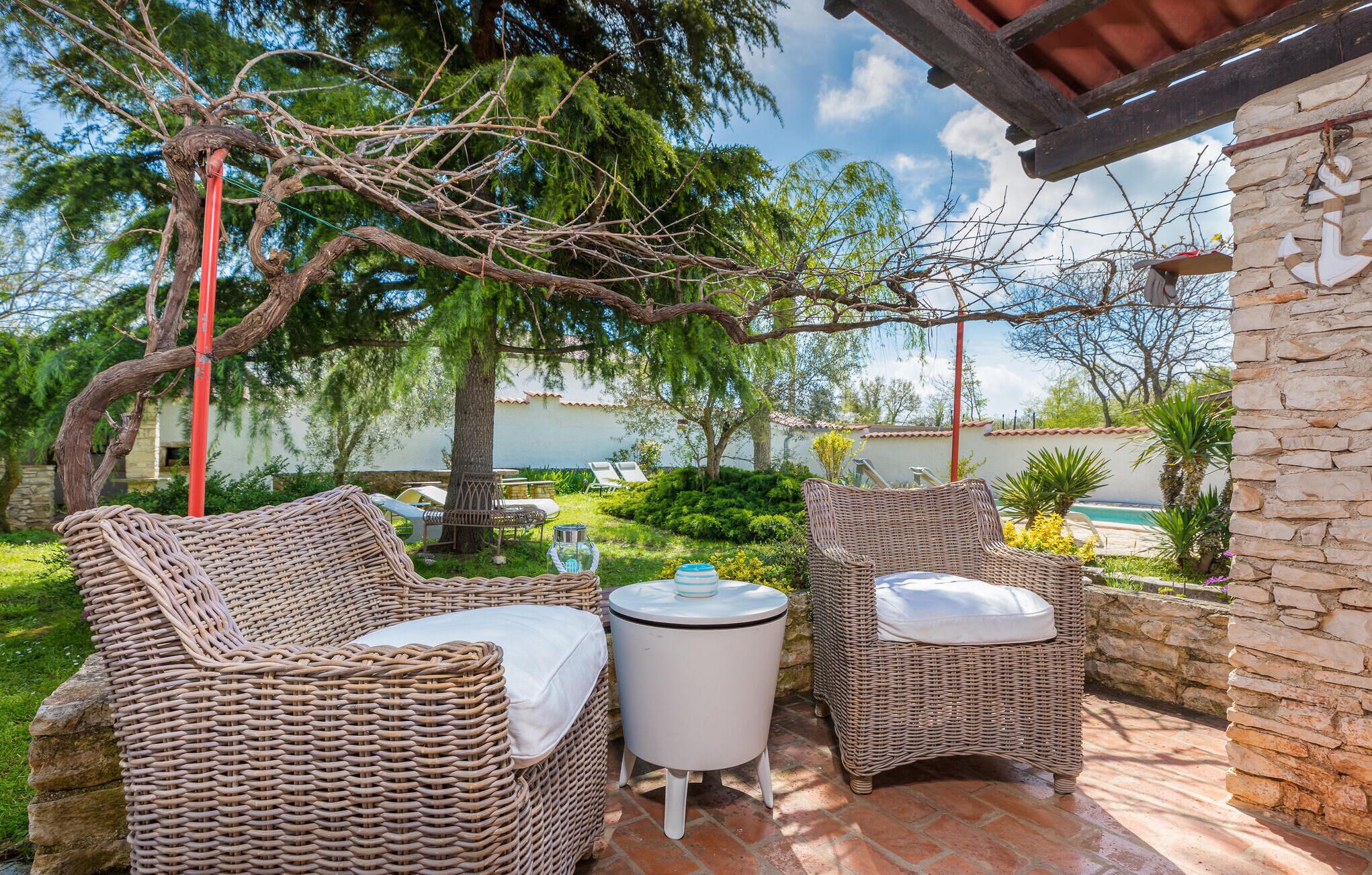A patio area with wicker furniture and a small table, surrounded by greenery and a stone wall.