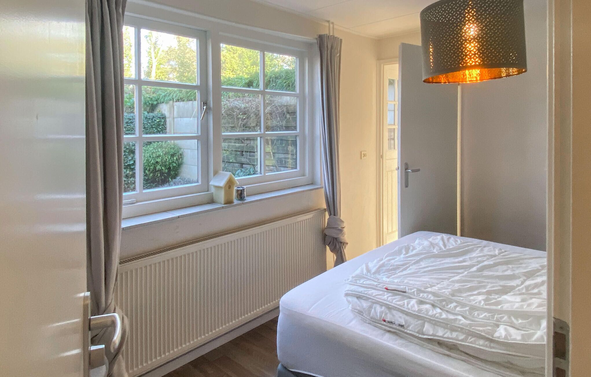 Une chambre à coucher avec un lit, un radiateur, une fenêtre avec des rideaux et une lampe suspendue.