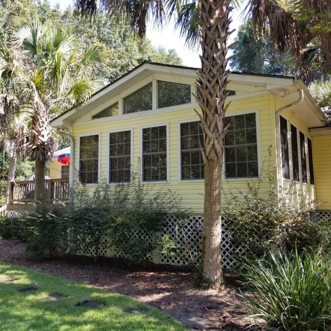 Une maison jaune avec de nombreuses fenêtres, entourée de verdure et d’une clôture en bois.