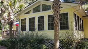 A yellow house with multiple windows, surrounded by greenery and a wooden fence.