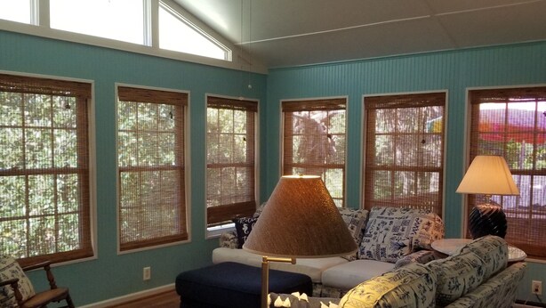 A living room with a blue sofa, a blue ottoman, a patterned armchair, and a floor lamp. There are large windows with blinds, a ceiling fan, and a rug on the floor.