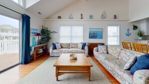 Una sala de estar con una mesa de centro de madera, sofás con estampado floral, una zona de comedor con sillas de madera y vistas a un edificio a través de un ventanal grande.