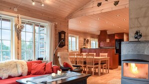 A cozy cabin interior with a fireplace, wooden furniture, and a large window overlooking a forest.