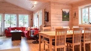 A cozy cabin interior with a wooden dining table, chairs, a red sofa, and a large window with curtains.