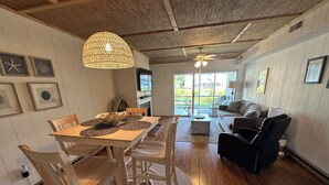 Un coin repas avec une table en bois, des chaises et un élément décoratif central en fibres tressées. La pièce adjacente, qui sert de salon, est équipée d’un ventilateur de plafond, d’une télévision et d’un canapé.