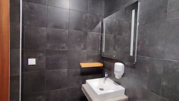 Ein modernes Badezimmer mit dunklen Fliesen, einem weißen Waschbecken, einem Seifenspender und einem Holzregal.