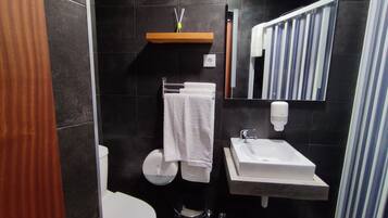 Un baño moderno con inodoro, lavamanos y espejo. Hay un perchero con una toalla, un dispensador de jabón y un pequeño estante con una botella.